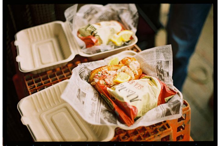 Takeout trays with lobster rolls, lemon slices, and chips on a crate.