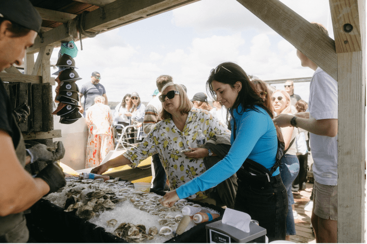 People gathering at an outdoor oyster bar, selecting oysters from a tray.