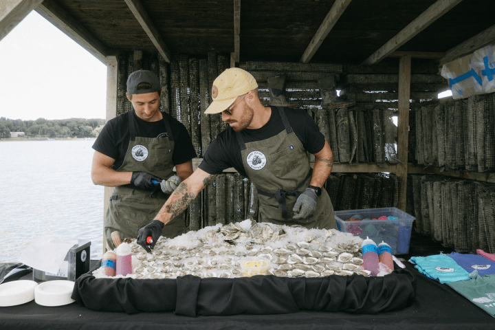 Two workers wearing aprons shucking oysters on a covered pier by a body of water.